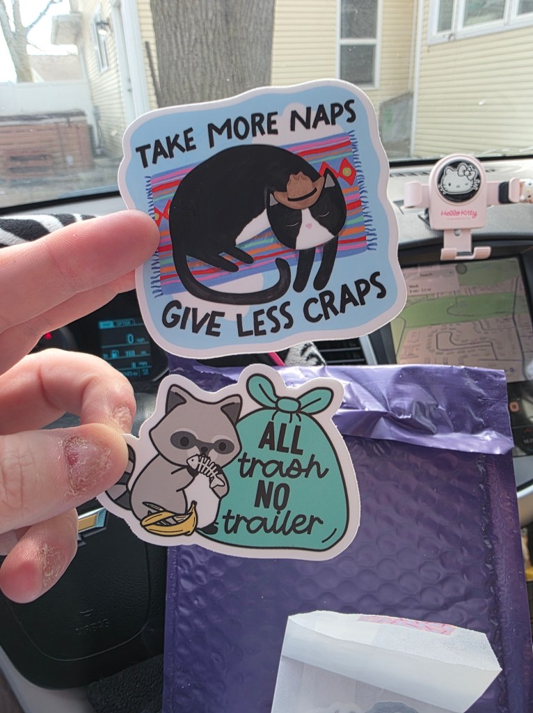 A hand holding two stickers. The first has a cat sleeping and says, "Take more naps give less craps." The other is a raccoon next to a trash bag & eating garbage, with the words "All Trash No Trailer" written on the bag.