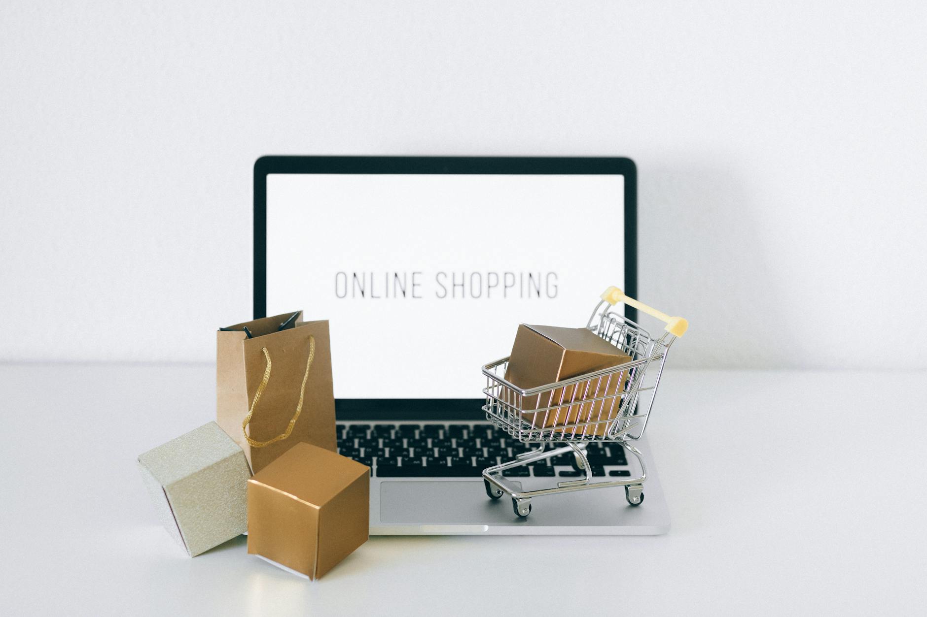 Laptop with the words "ONLINE SHOPPING" written in caps and a display of two tiny boxes, a bag, and a mini shopping cart with a box inside.