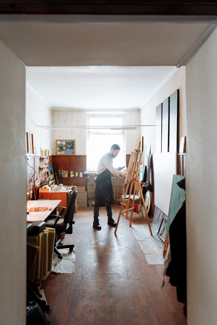 Artist at an easel, painting. He's standing in a room cluttered with art, tables, chairs, and painting supplies.