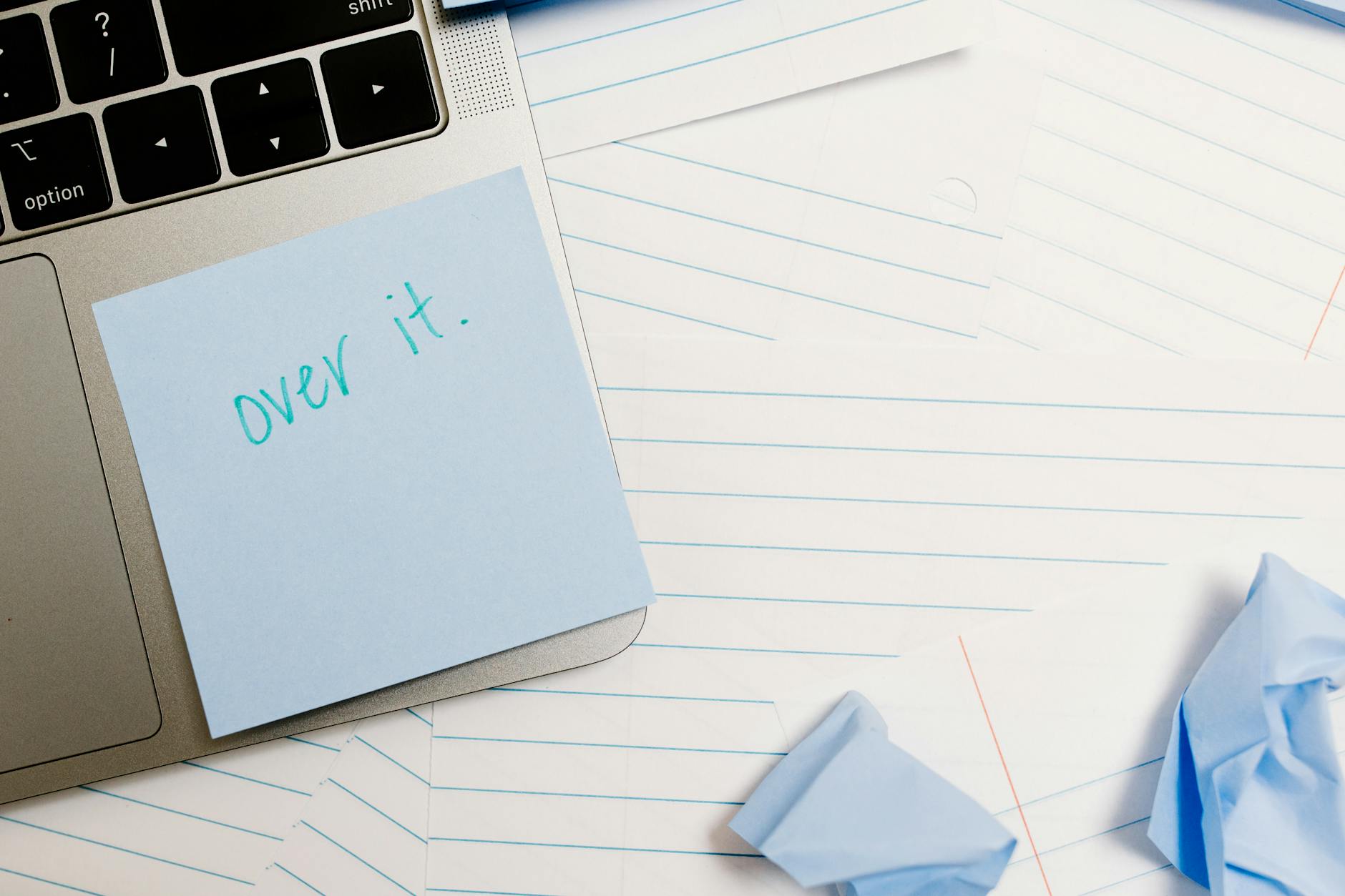 a background with a laptop, lined paper, and crunched up post it notes, with a post it stuck to the corner of the laptop that says, "Over it."